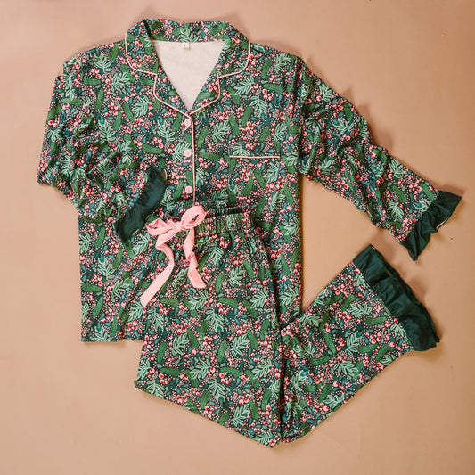 The Jolly Sprig Sweet Dreams Pajama Pants Set by The Darling Effect features a green background adorned with a festive holly and berry pattern, accented with pink piping, a matching bow, and ruffled cuffs on both the sleeves and pants. Crafted from ultra-soft fabric for ultimate comfort, this cozy pajama set is displayed laid flat against a neutral background.