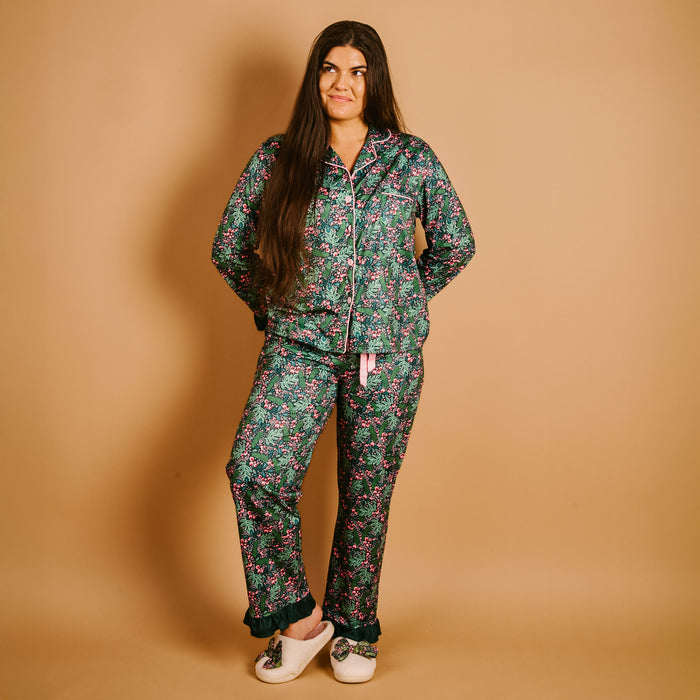 Wearing The Darling Effect’s Jolly Sprig Sweet Dreams Pajama Pants Set, a person with long brown hair stands in ultra-soft floral pajamas and slippers against a plain tan background, hands behind their back, smiling slightly while looking off to the side.