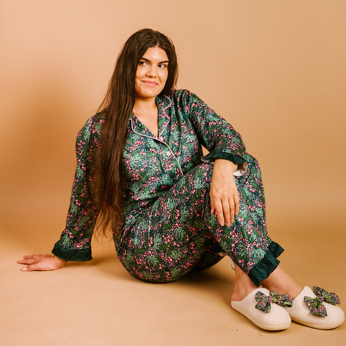 A woman with long dark hair sits on the floor, smiling, wearing The Darling Effect’s Jolly Sprig Sweet Dreams Pajama Pants Set crafted from ultra-soft fabric in a floral green design, paired with cream slippers adorned with bows, set against a beige background.