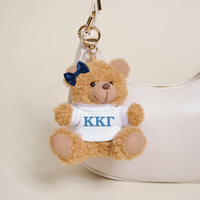 The Darling Effect's Kappa Kappa Gamma Teddy Bear Bag Charm features a navy bow and white shirt with blue ΚΚΓ letters, hanging from a white handbag—an adorable Bid Day gift for any sorority member.