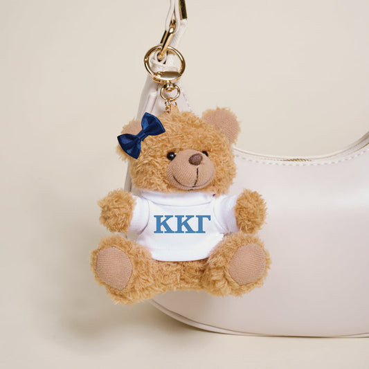 The Darling Effect's Kappa Kappa Gamma Teddy Bear Bag Charm features a navy bow and white shirt with blue ΚΚΓ letters, hanging from a white handbag—an adorable Bid Day gift for any sorority member.