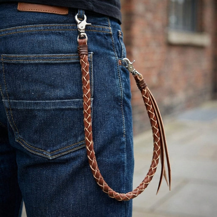 Light brown braided leather wallet chain with silver lobster claw clasps attached to dark blue denim jeans back pocket, featuring decorative leather fringe tassels in urban street style setting