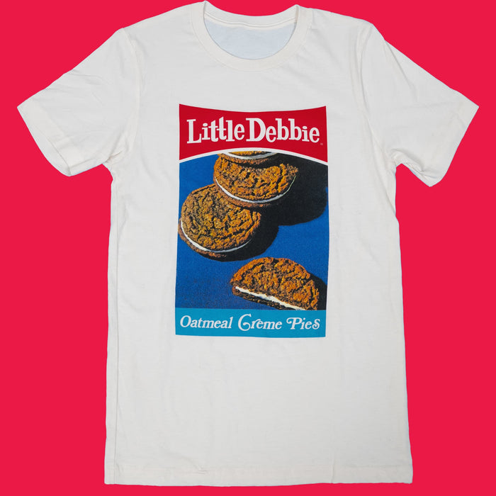 A white T-shirt featuring a retro design of Little Debbie Oatmeal Creme Pies. The shirt displays three oatmeal creme pies against a blue background with the "Little Debbie" logo in red at the top and "Oatmeal Creme Pies" in white text on a blue stripe at the bottom.