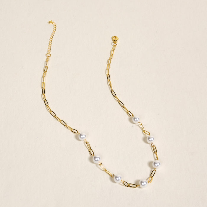 The Darling Effect Pearl Paperclip Necklace Chain, featuring elongated links, spaced white pearls, and delicate charms, is displayed on a light beige surface.