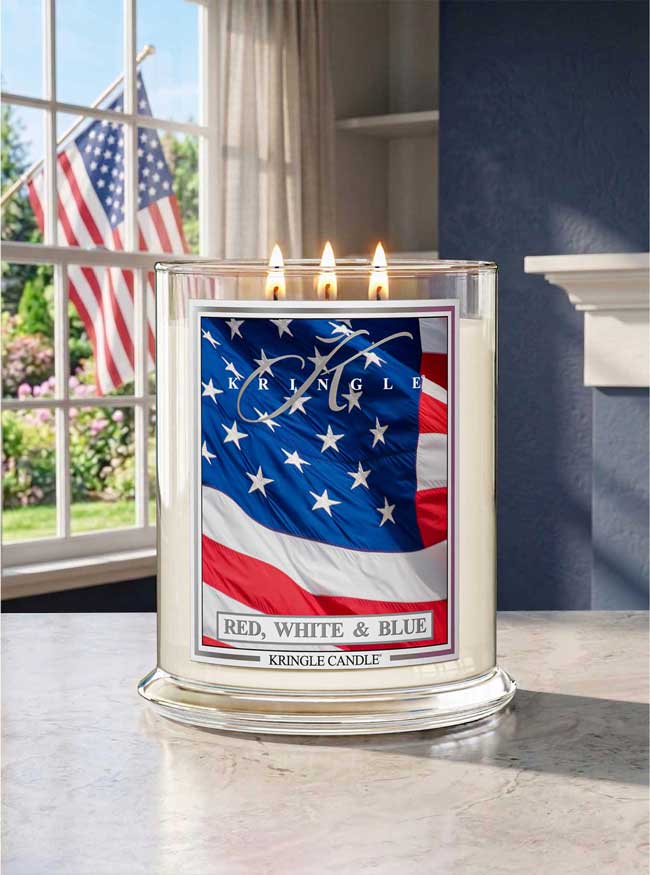 Kringle Candle Red, White & Blue candle with American flag design on a windowsill with an American flag outside.