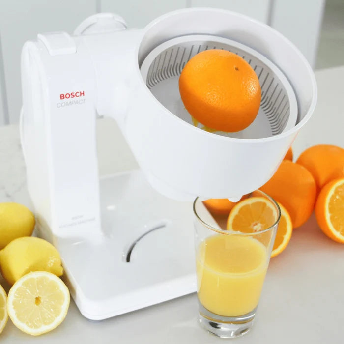 Bosch compact mixer with citrus juicer attachment juicing an orange next to a glass of orange juice and fresh citrus on a white kitchen counter