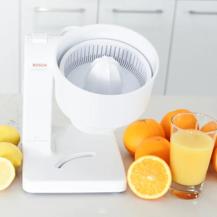 Bosch compact mixer with citrus juicer attachment on a white kitchen counter next to lemons, oranges, and a glass of orange juice