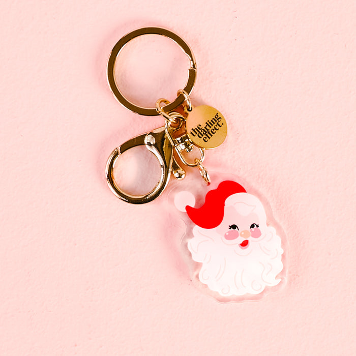 The Darling Effect’s Santa Acrylic Keychain features a cute, cartoon-style Santa face charm with a red hat on a gold clip and round tag. Made from durable acrylic, it’s shown on a light pink background.