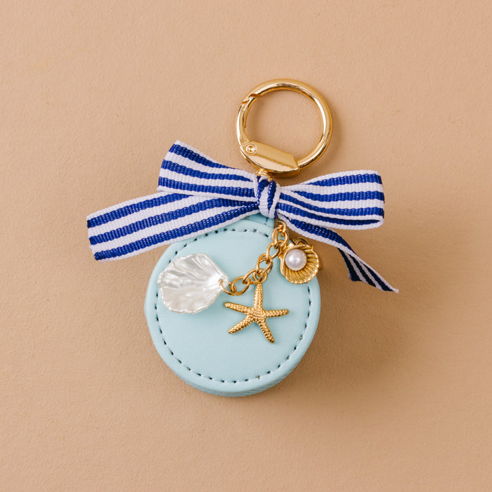 The Darling Effect Sea Breeze Travel Jewelry Case Bag Charm: a round, light blue keychain with gold ring, striped bow, gold starfish and shell charms, faux pearl, and clear shell charm. Photographed on beige.