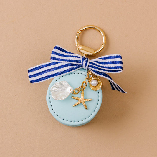 The Darling Effect Sea Breeze Travel Jewelry Case Bag Charm: a round, light blue keychain with gold ring, striped bow, gold starfish and shell charms, faux pearl, and clear shell charm. Photographed on beige.