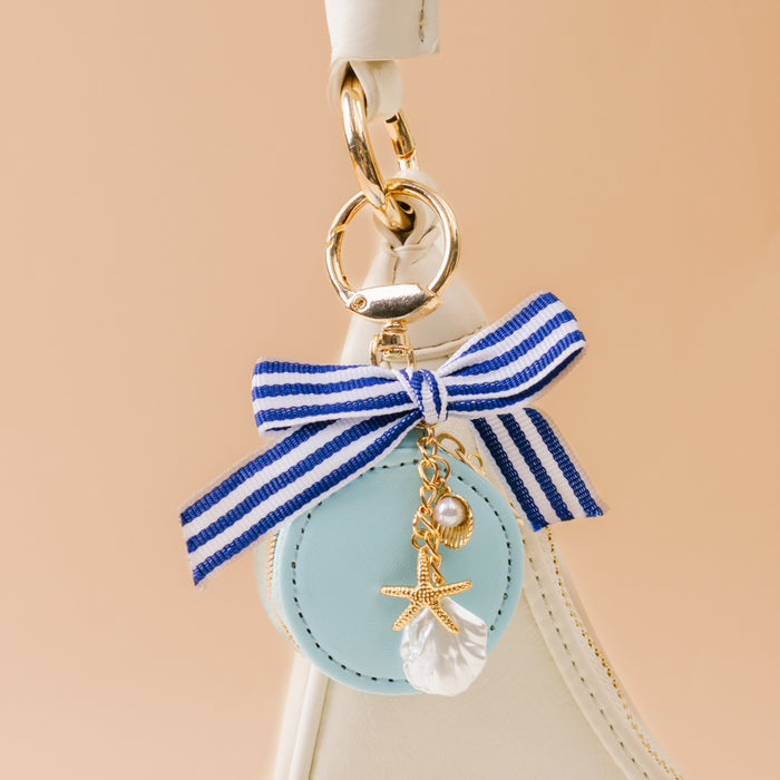 A close-up of The Darling Effect's Sea Breeze Travel Jewelry Case Bag Charm with a blue and white striped bow, gold starfish, pearl, seashell, and light blue charm—perfect for accessorizing your portable jewelry organizer.