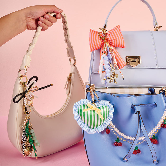 Three handbags from The Darling Effect, featuring Sea Breeze Travel Jewelry Case Bag Charms, ornate keychains, and ribbons, are displayed on a pastel pink background. A hand holds a cream bag while two blue ones are nearby.