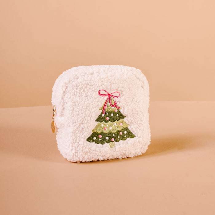 The Square Twinkling Tree Teddy Pouch by The Darling Effect is a white, fluffy sherpa pouch adorned with an embroidered green Christmas tree decorated with pink ornaments and topped with a pink bow, all set against a light beige background—making it the perfect festive holiday accessory for the season.