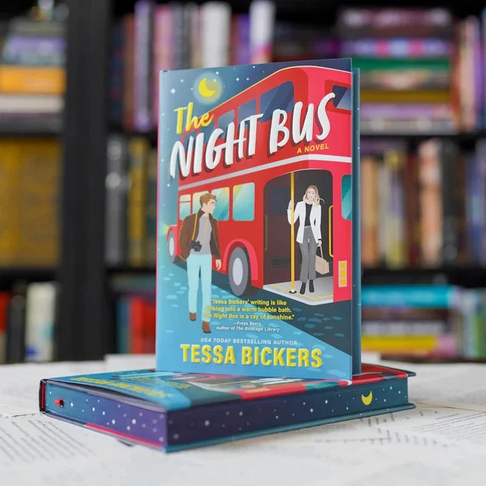 The Night Bus paperback edition by USA Today bestselling author Tessa Bickers standing upright, showing the original publisher cover with illustrated characters boarding a red double-decker London bus under a crescent moon, stacked on top of The Night Bus LitJoy Crate Edition hardcover, set against a blurred bookshelf background.