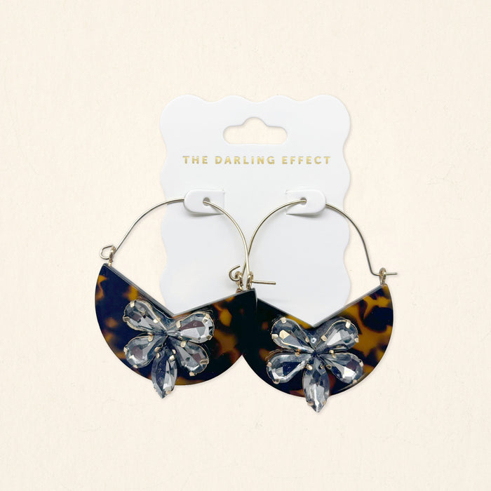 The Darling Effect’s Totally Tortoise Marble Bloom Hoop Earrings feature half-moon tortoiseshell pendants and crystal floral accents, displayed on a branded white card.