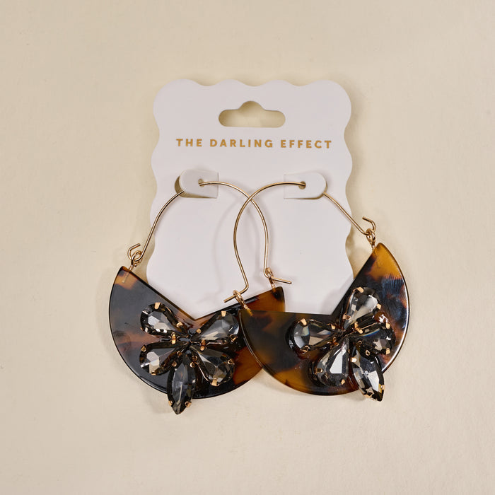 The Darling Effect’s Totally Tortoise Marble Bloom Hoop Earrings feature large gold hoops with brown tortoiseshell crescents and rhinestone butterfly accents—stylish, modern statement pieces on a white branded card.