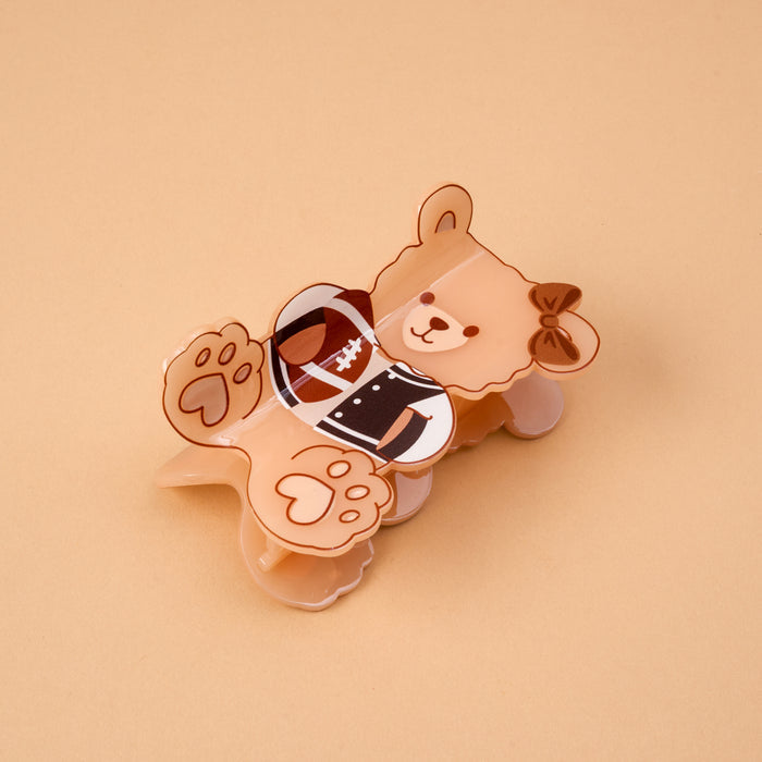 The Touchdown Teddy Shape Up Claw Clip by The Darling Effect is a charming hair accessory shaped like a brown bear in a football uniform, complete with a bow on its head and its paws visible. Shown lying on a beige background, this cute claw clip is perfect for adding an adorable touch to your hairstyle.