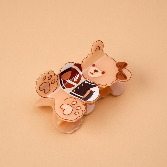 The Touchdown Teddy Shape Up Claw Clip by The Darling Effect is a charming hair accessory shaped like a brown bear in a football uniform, complete with a bow on its head and its paws visible. Shown lying on a beige background, this cute claw clip is perfect for adding an adorable touch to your hairstyle.