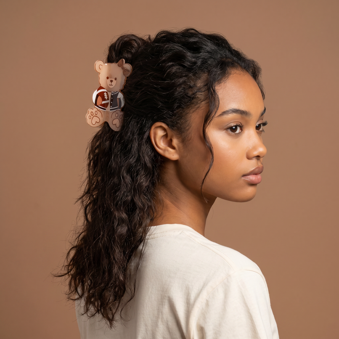 A woman with wavy dark hair, styled in place with The Darling Effect's Touchdown Teddy Shape Up Claw Clip, stands confidently against a plain brown background, looking off to the side. She wears a white shirt and her cute teddy bear hair accessory adds a playful touch to her look.