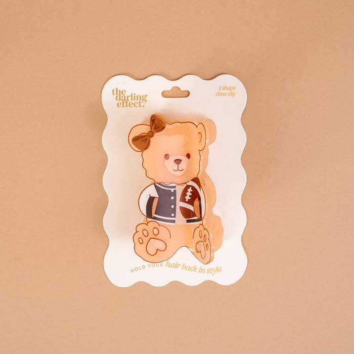 Add a sporty touch to your hairstyle with the Touchdown Teddy Shape Up Claw Clip by The Darling Effect—a teddy bear-shaped hair accessory featuring a brown bow, packaged on a white wavy-edged card with "The Darling Effect" and "Hold your hair back in style." The adorable bear illustration wears a letterman jacket and holds a football, making this claw clip perfect for cute, game-day looks.