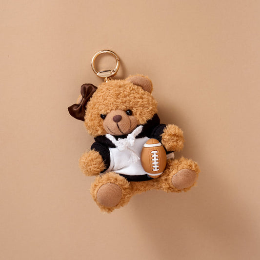 The Touchdown Teddy Teddy Bear Bag Charm by The Darling Effect features a small teddy bear keychain dressed in a black and white hoodie and holding a tiny plush football, displayed on a beige background—a delightful accessory for your bag and an ideal choice for fans of charming plush charms.