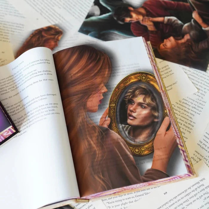 We Who Will Die LitJoy Edition page overlay inside an open book, depicting a woman with long auburn hair gazing at a gold-framed oval mirror reflecting a young man's face, with other overlays and the book in the background