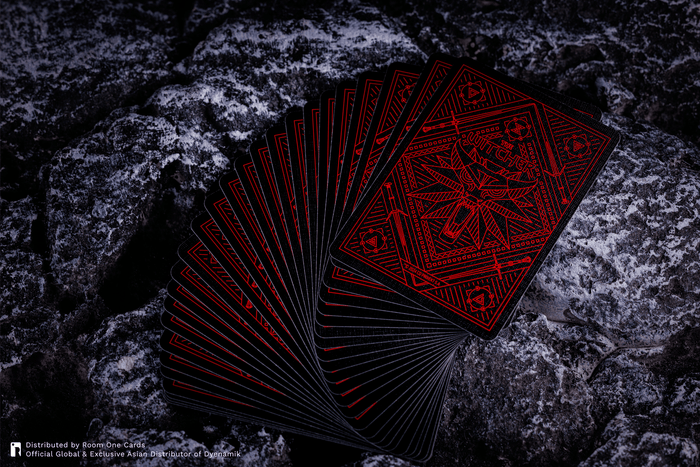A fan of black playing cards with intricate red occult-style geometric designs, displayed on a dark, textured rock surface. The Witcher Playing Cards by Room One.