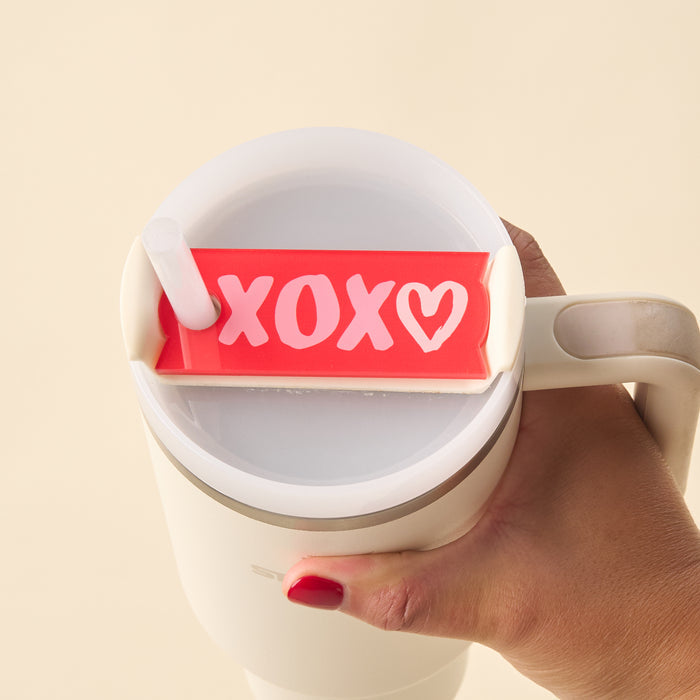 A hand with red-painted nails holds a white travel mug topped with The Darling Effect’s XOXO Tumbler Lid Tag—a red Quencher 2.0 accessory featuring “XOXO” and a heart in white, adding a cute personalized touch.